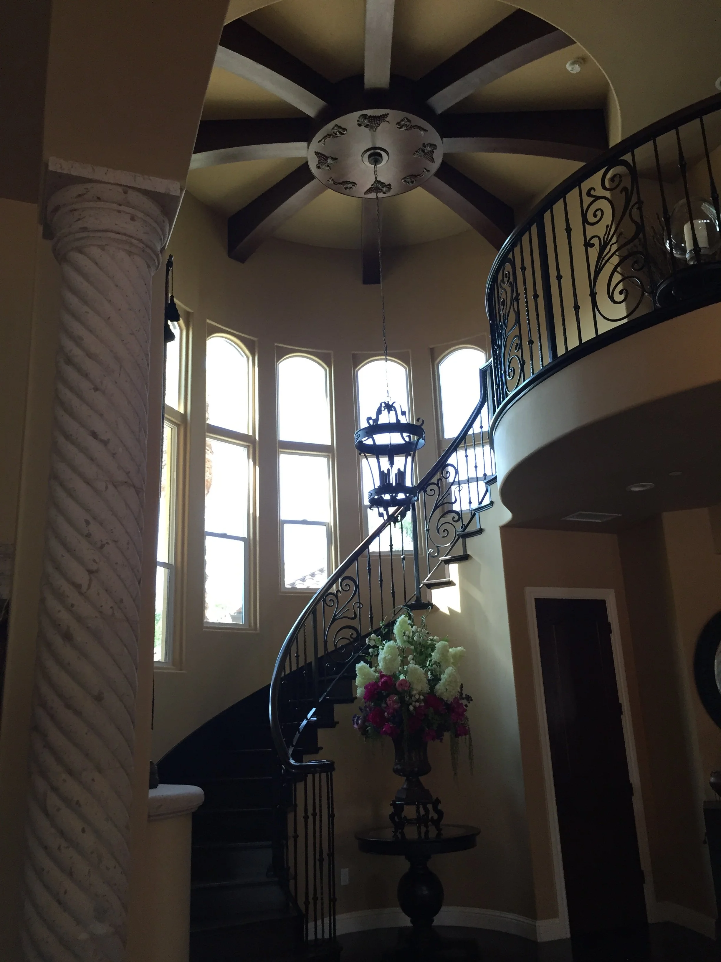 Interior view of a spiral staircase with ornate black iron railings, a bouquet of white and pink flowers on a black pedestal table, tall curved windows allowing natural light, and a ceiling with exposed wooden beams and a decorative chandelier.