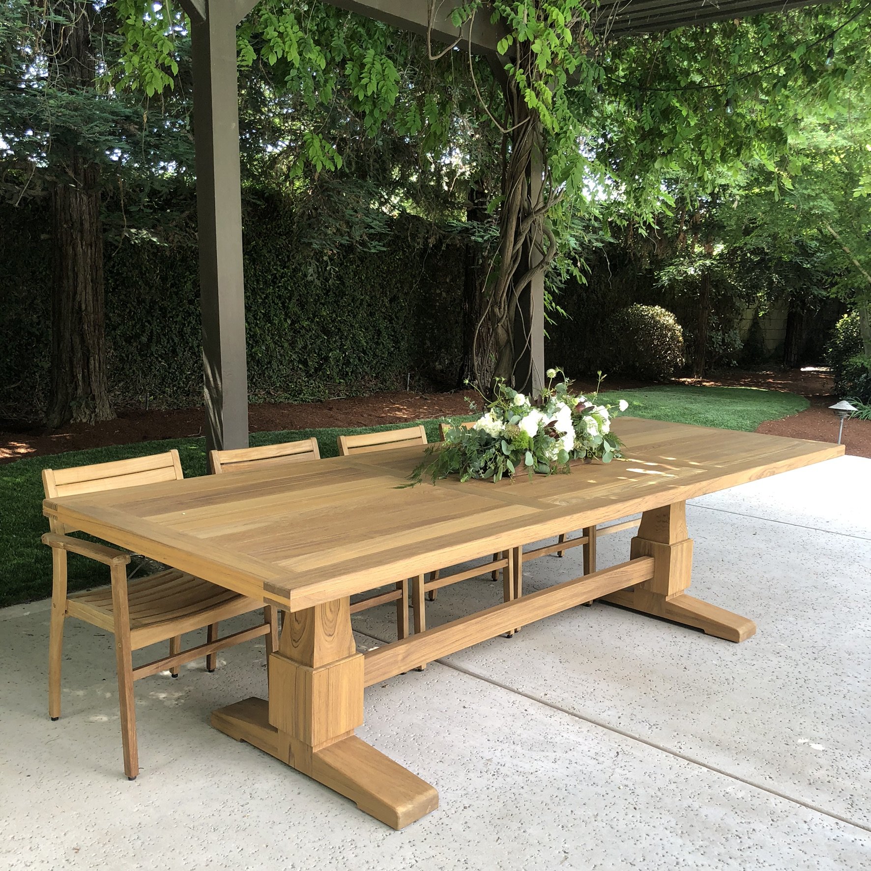 Outdoor wooden dining table with a floral centerpiece on a patio, shaded by greenery.