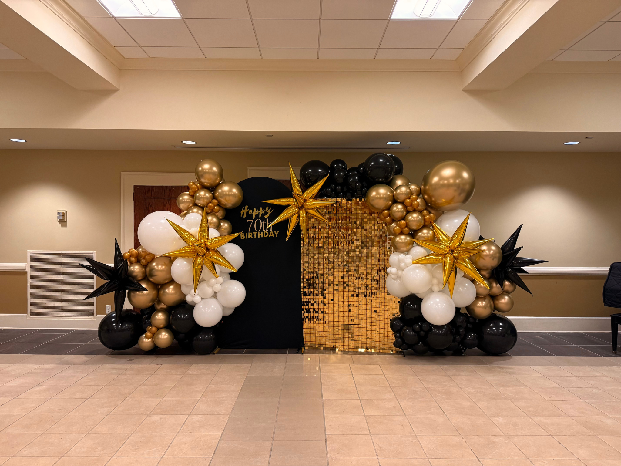 Decorative balloon arch with black, white, gold, and black star-shaped balloons, set against a gold sequin backdrop. A black circle with gold lettering reads "Happy 70th Birthday."