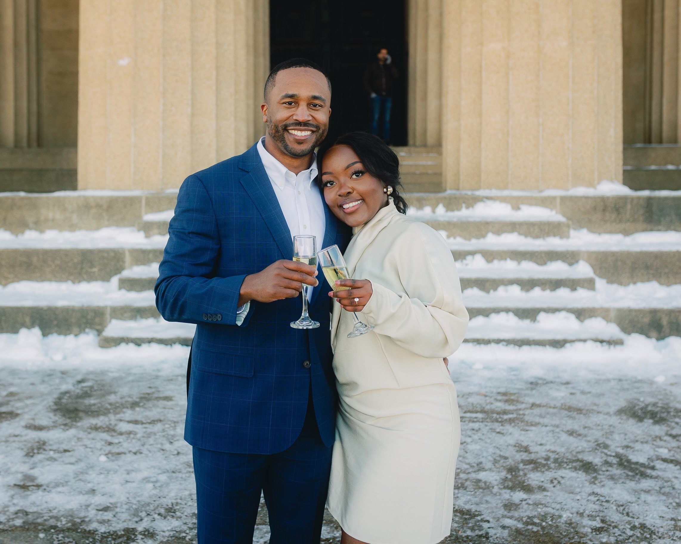 A smiling couple dressed in formal attire standing outside on snow, holding champagne glasses, in front of a building with steps.