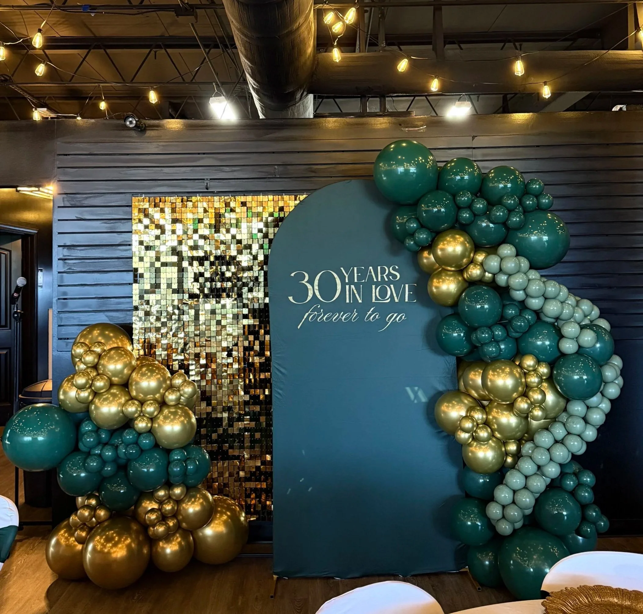 Decorative backdrop celebrating 30 years in love with gold and teal balloons arranged around a teal sign that reads "30 Years in Love forever to go". The backdrop includes a black wall with string lights and a mirrored mosaic panel.