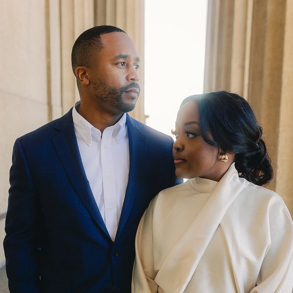 A man and woman standing indoors, looking at each other. The man has short hair, a beard, and is wearing a dark blue suit with a white shirt. The woman has curly black hair, gold earrings, and is wearing a white outfit.