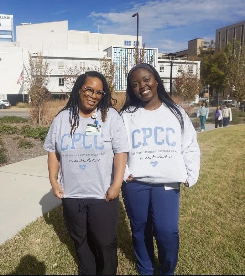 Two women standing outdoors on a sunny day, wearing matching gray sweatshirts with blue lettering that reads 'CPCC' and 'nurse', smiling at the camera with a modern cityscape and other people in the background.