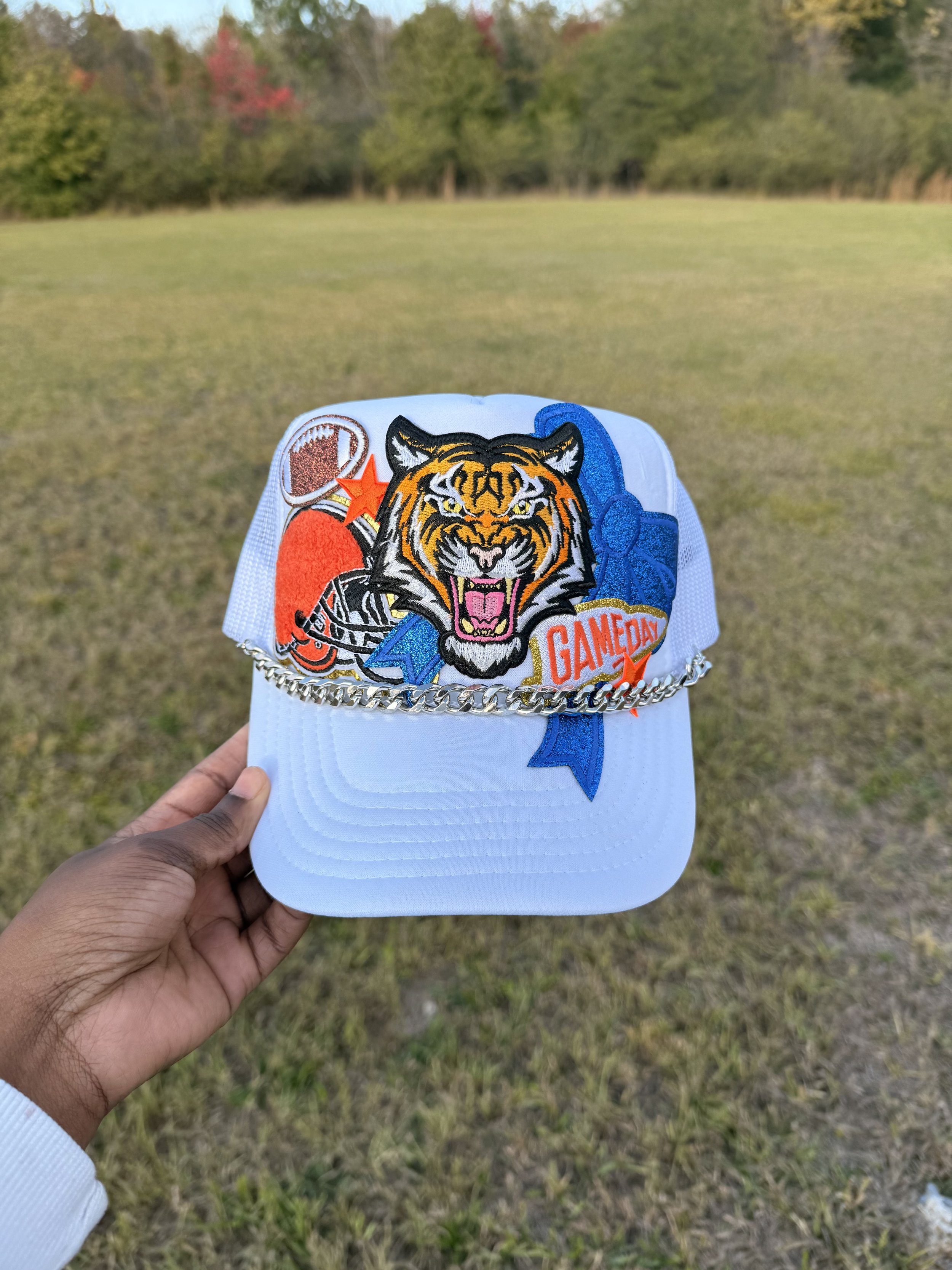 A white baseball cap with colorful patches of a football, a football helmet, a roaring tiger, and the word "GAMEDAY". There is a silver chain around the cap's brim. The background is an empty grassy field with trees in fall colors.