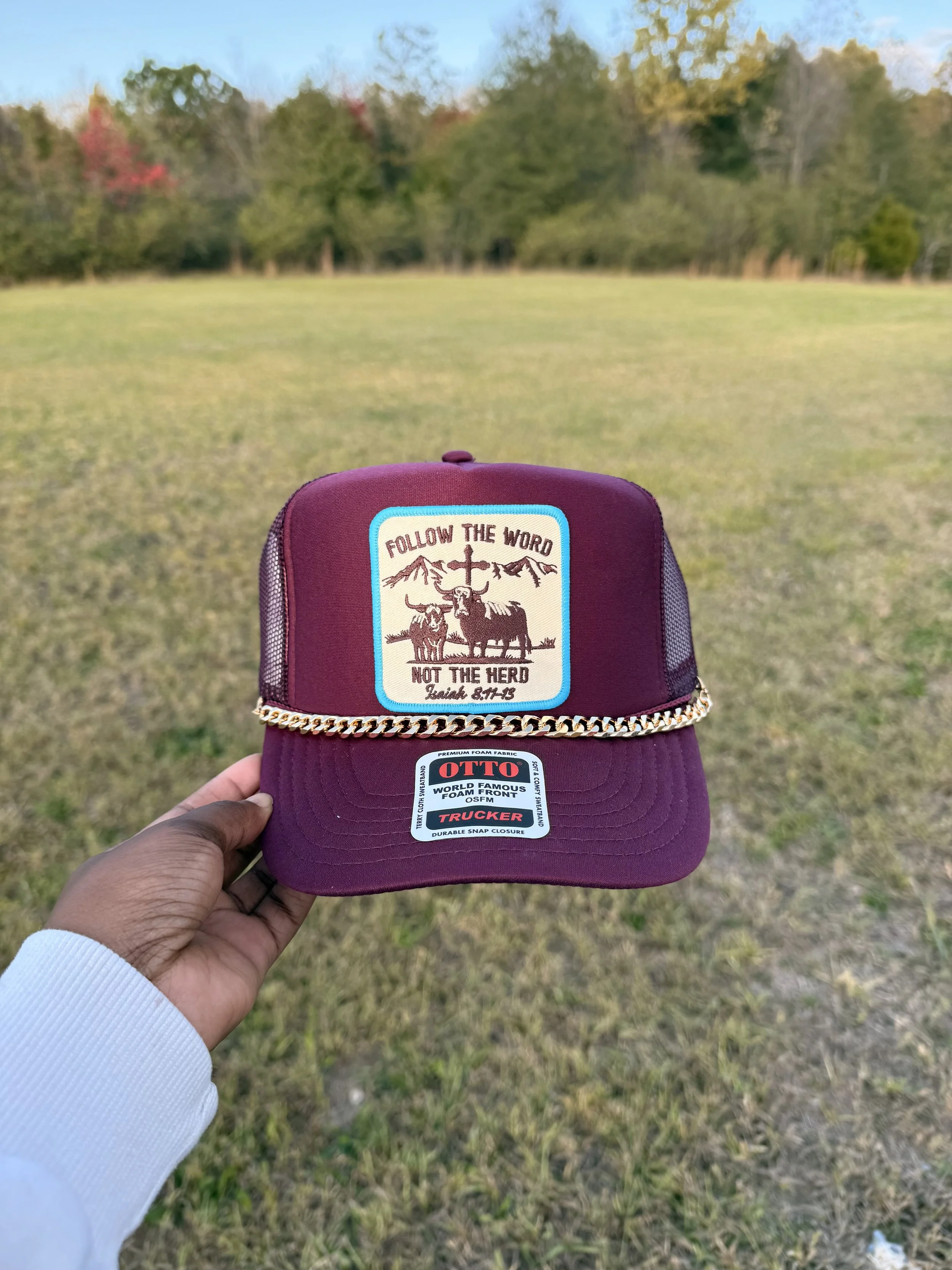 A person holding a maroon baseball cap with patches in an open grassy field with trees in the background. The cap has a patch with the text 'Follow the Word Not the Herd,' an illustration of cattle and mountains, and a cross. Another sticker on the cap reads 'OTTO' and 'Trucker'.