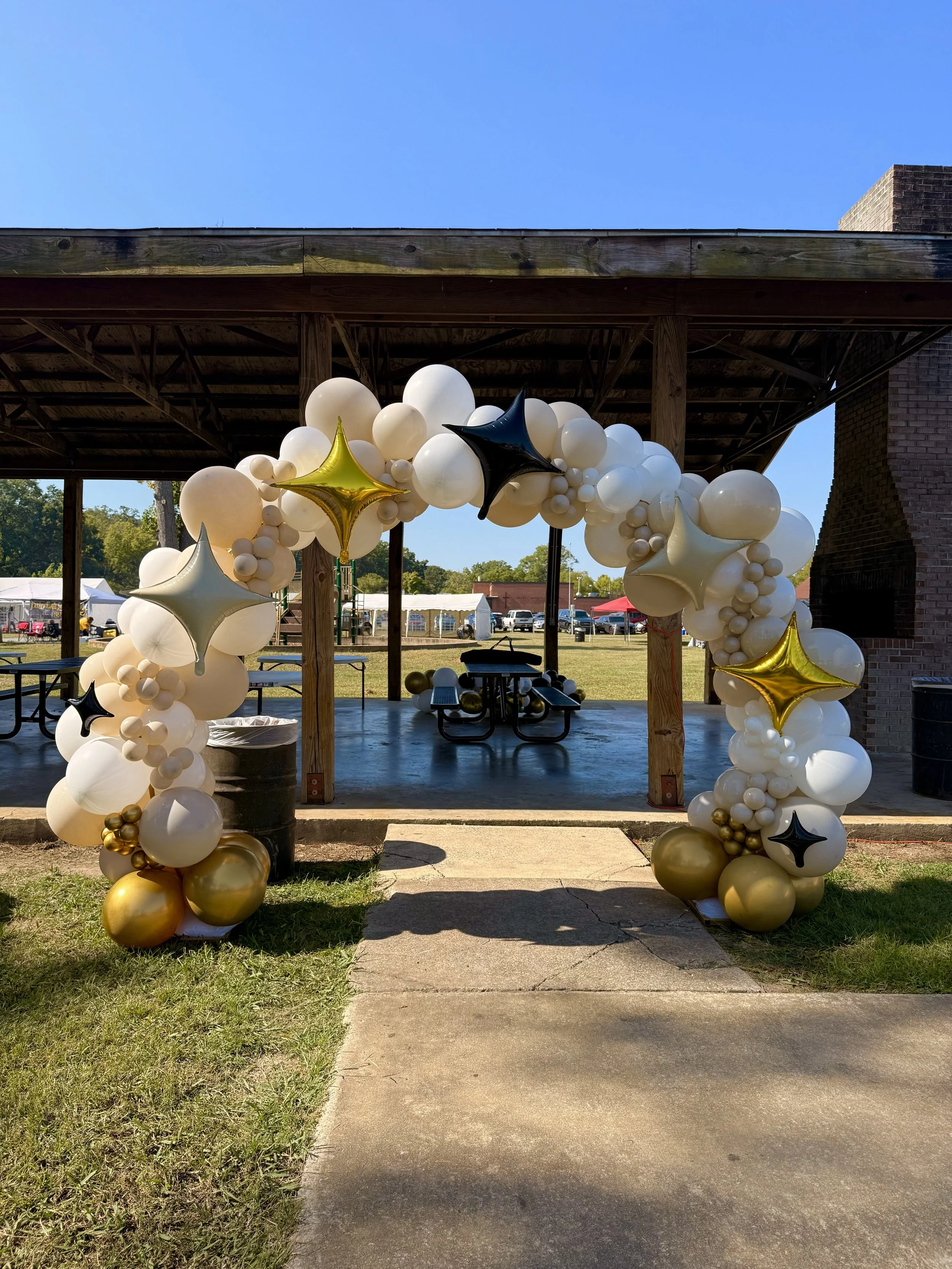 Balloon arch with white, black, gold, and silver balloons at an outdoor event under a pavilion.