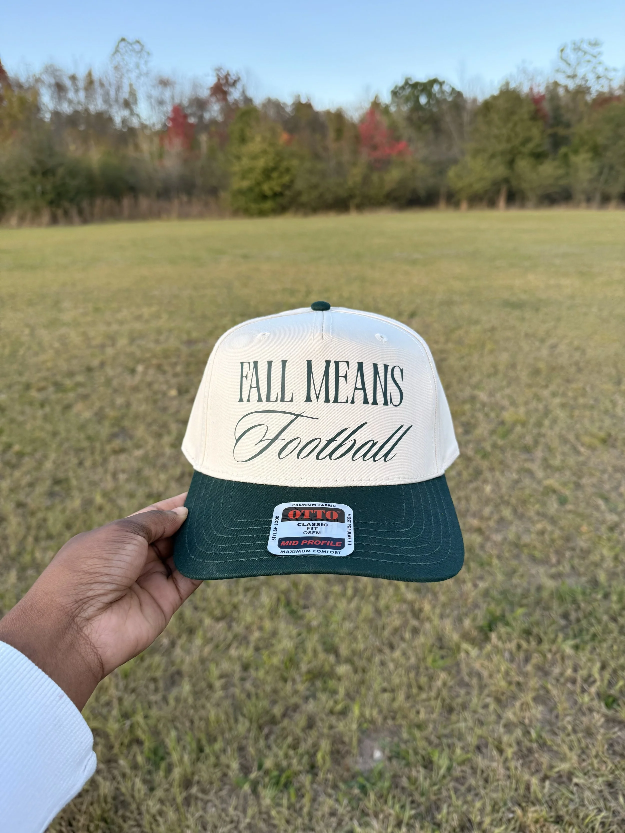 Hand holding a hat with a white front and black brim, with the text 'Fall Means Football' embroidered on the front, set against an open grassy field with trees in autumn colors in the background.