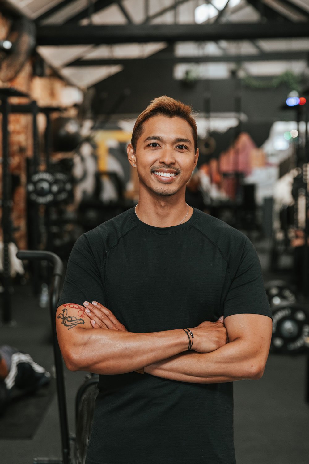 Smiling man with arms crossed, in a gym with workout equipment in the background.