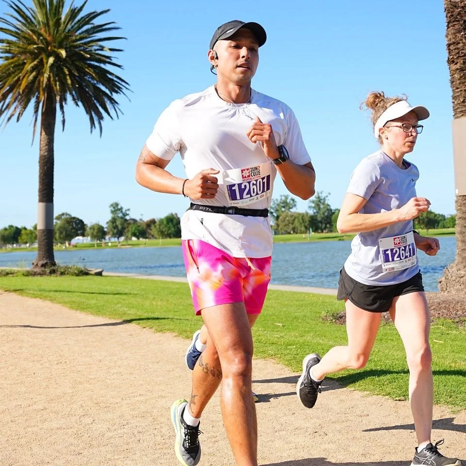 Two runners participating in a race along a park path near a lake, under a clear blue sky, with palm trees and greenery in the background.