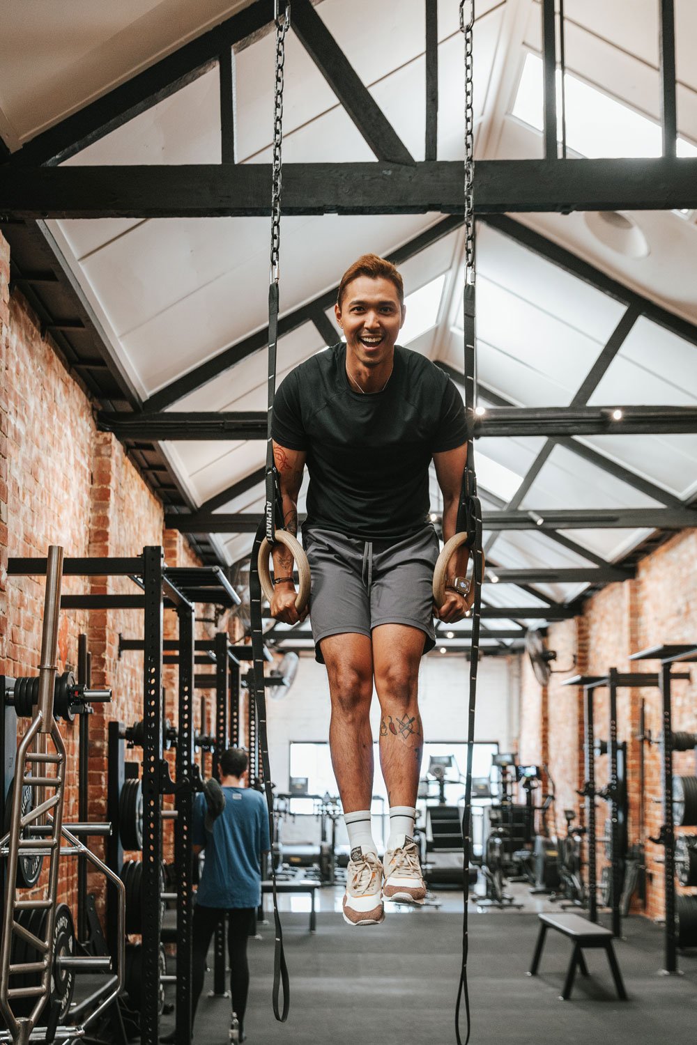 A man smiling while doing exercises with gymnastic rings in a fitness gym with brick walls and high ceilings.