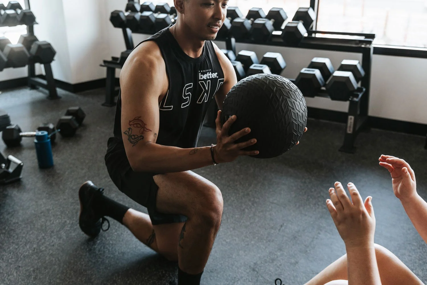 A man kneeling on one knee, holding a medicine ball during a workout in a gym. There are dumbbells and workout equipment in the background.