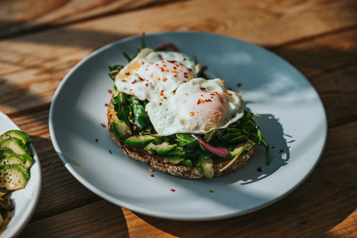 A plate with toasted bread topped with sliced avocado, fresh greens, and two poached eggs seasoned with red pepper flakes, on a wooden table.