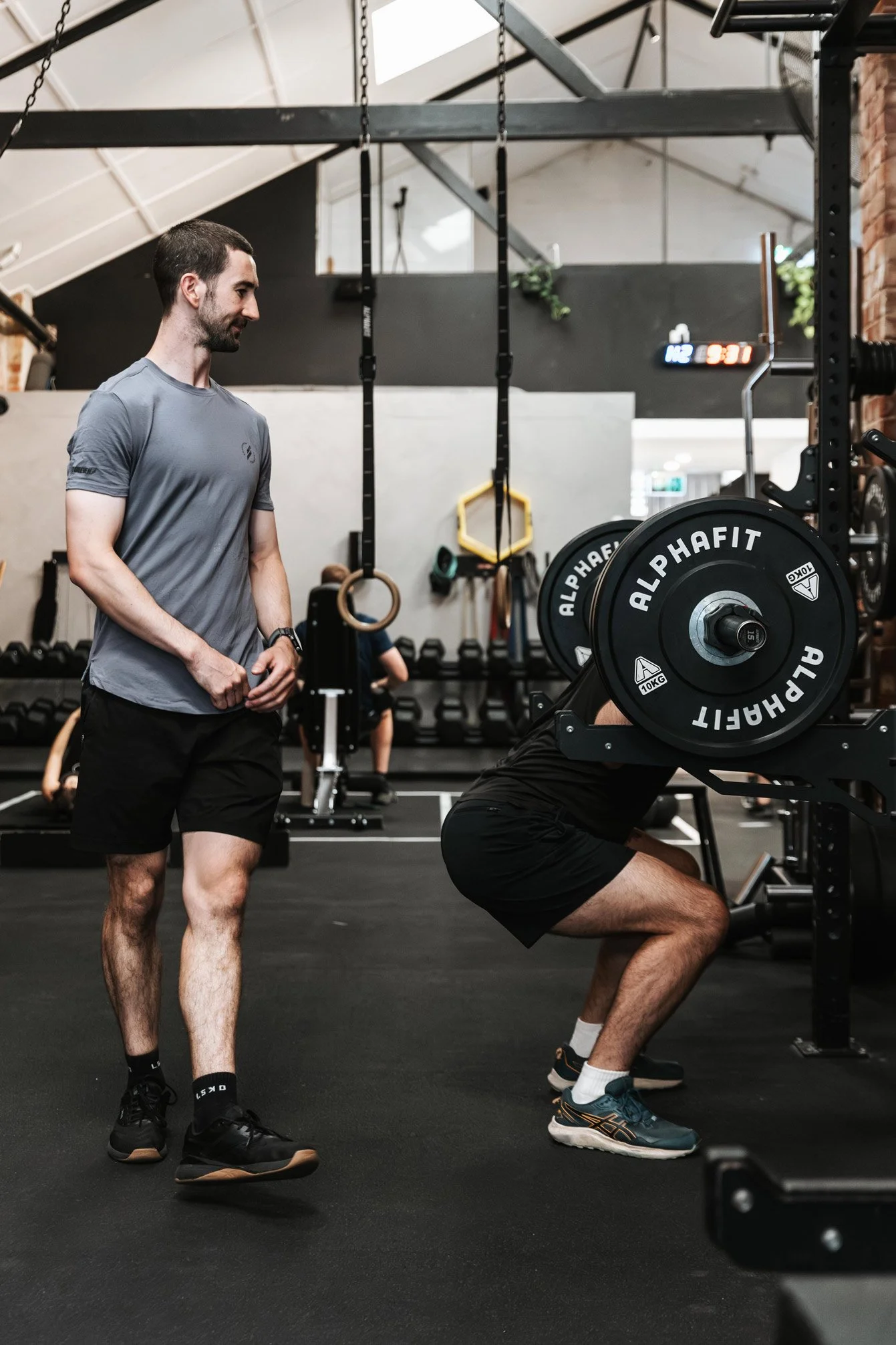A man is performing a squat exercise with a barbell on his shoulders, while another man stands nearby supervising in a gym.