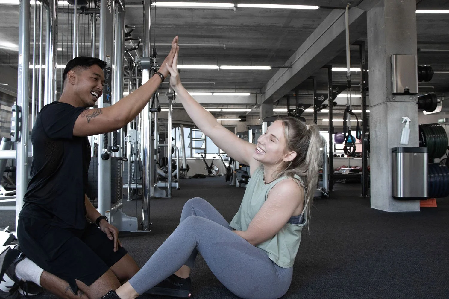 A woman and a man high-fiving at a gym, with gym equipment in the background.