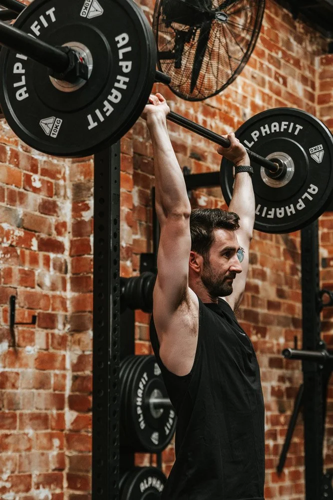 Man lifting a barbell overhead at a gym with brick wall background.