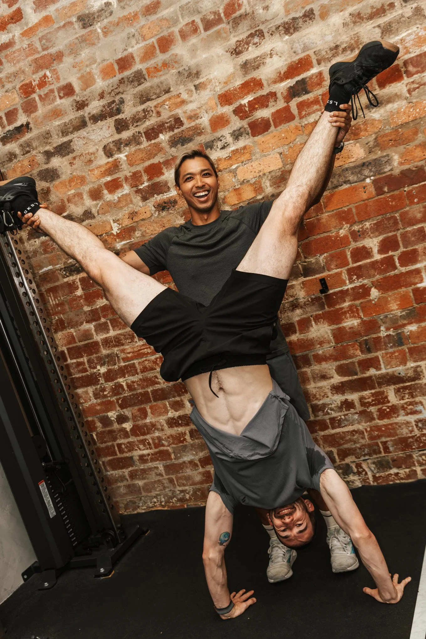Two men performing a handstand against a brick wall, with one man holding the other’s legs in a gym setting.