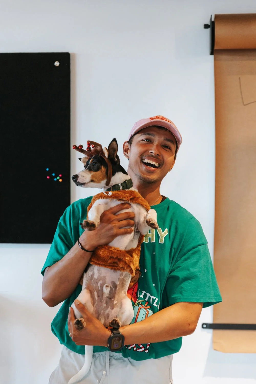 A man smiling and holding a dog dressed in a reindeer costume and antlers, indoors against a plain wall with a black bulletin board and a white wall.