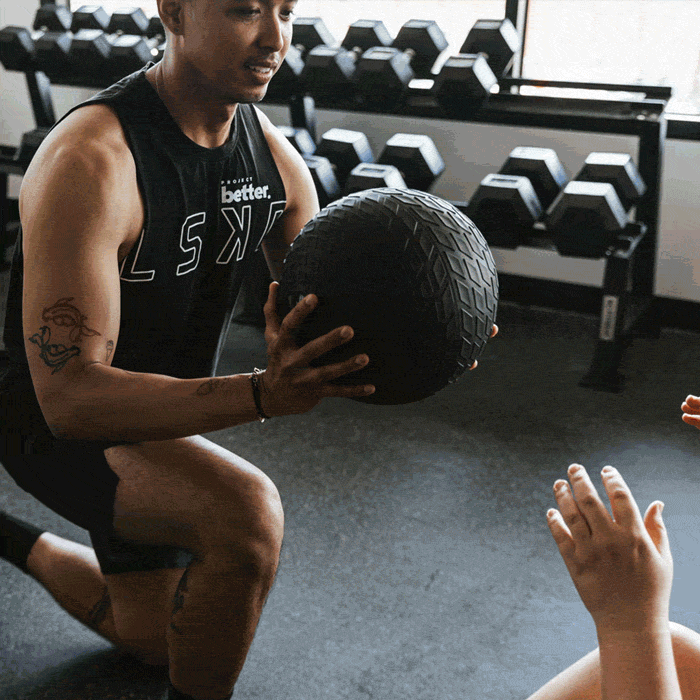 A person in a gym holding a medicine ball, with weights on racks in the background.