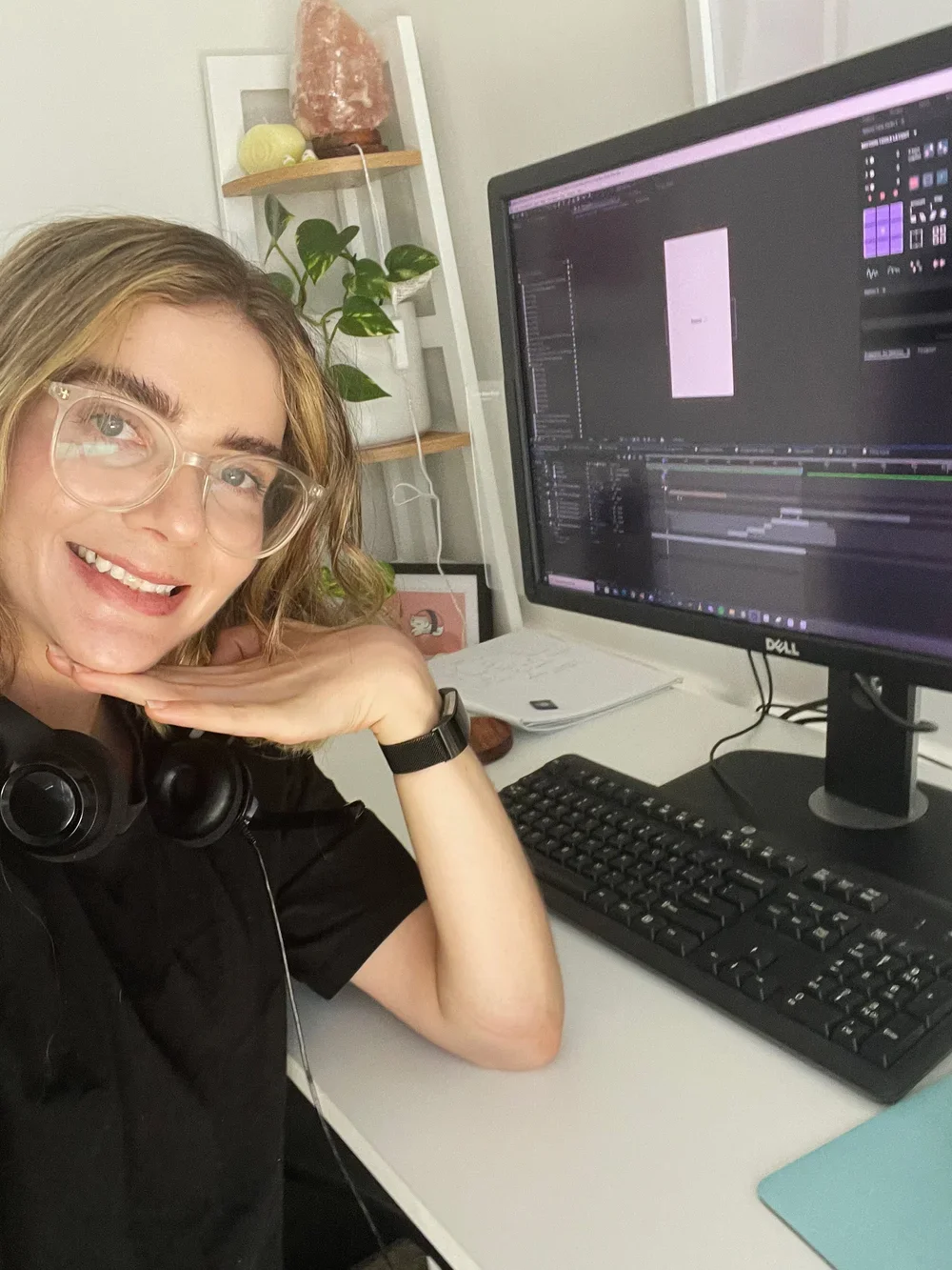 A woman with glasses smiling at the camera, sitting at a desk with a computer monitor displaying a video editing software, and a keyboard in front of her. There is a plant and salt lamp on a shelf in the background.