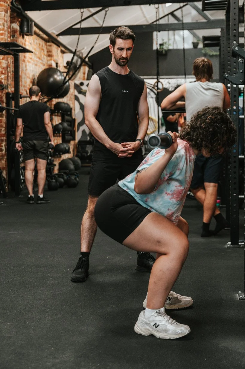 A woman performing a squat with a dumbbell at the gym, while a male trainer observes her technique.
