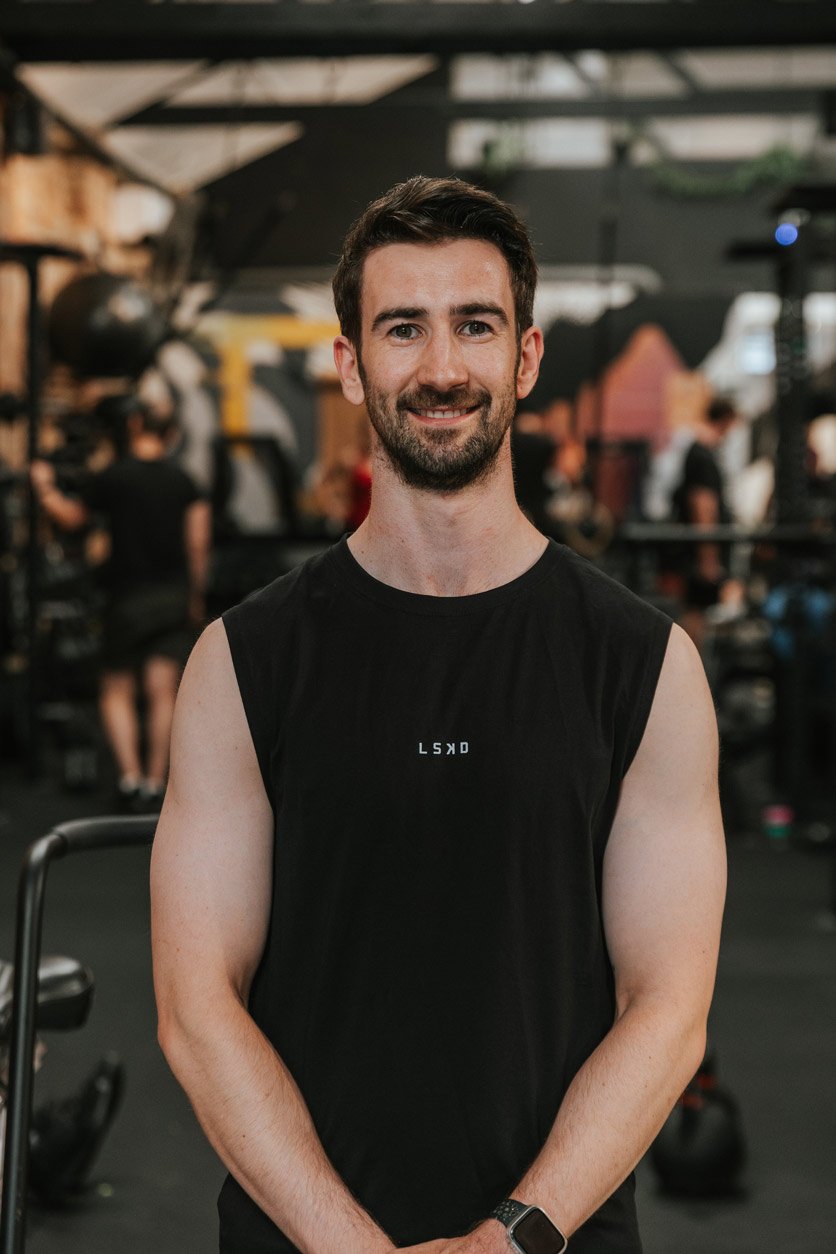 A young man with dark hair and a beard, smiling, in a gym. He is wearing a black sleeveless workout shirt and a smartwatch. The gym background shows other people working out and workout equipment.