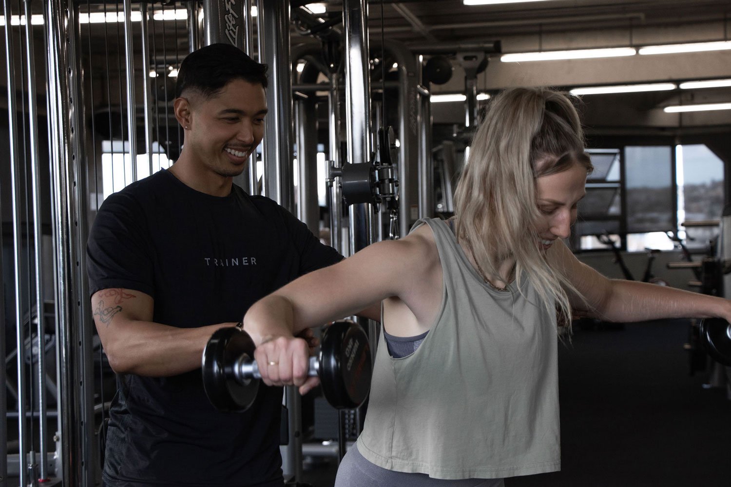 A female get instructions from a male trainer while lifting a dumbbell in a gym.