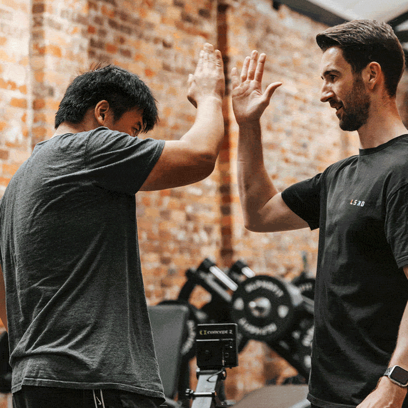 Two men giving each other a high five in a gym, with a brick wall background and workout equipment in the background.