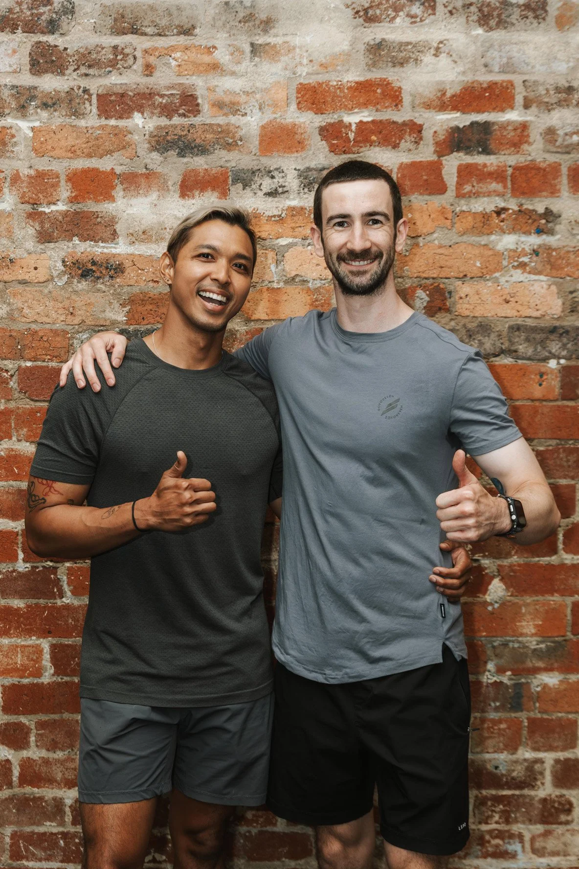 Two men smiling and giving thumbs-up, standing in front of a brick wall. One man has short blonde hair, tanned skin, and tattoos on his left arm. He is wearing a dark gray athletic shirt and gray shorts. The other man has short dark hair, a beard, light skin, and is wearing a light gray athletic shirt and black shorts. He is also wearing a smartwatch on his left wrist.