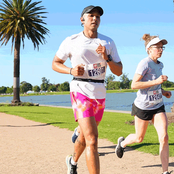 Two people running in a race near a lake on a sunny day, with trophy icons on their bibs and trees in the background.