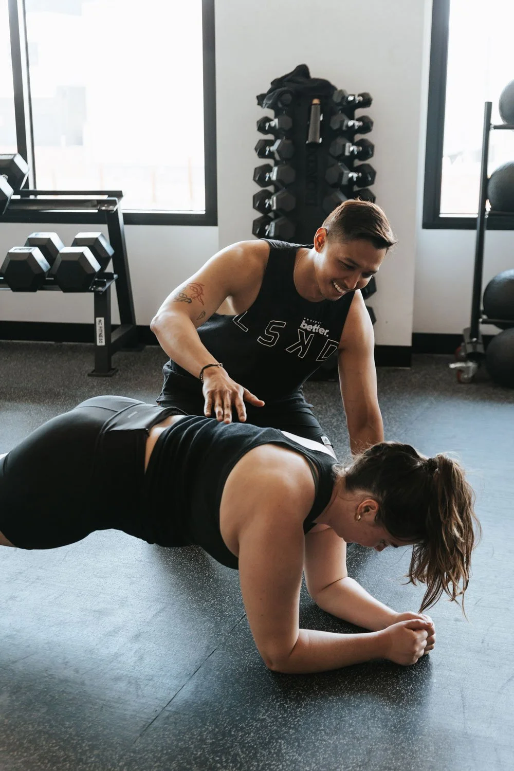 A woman doing a plank exercise on the floor with her forearms, while a fitness trainer watches and offers encouragement in a gym.