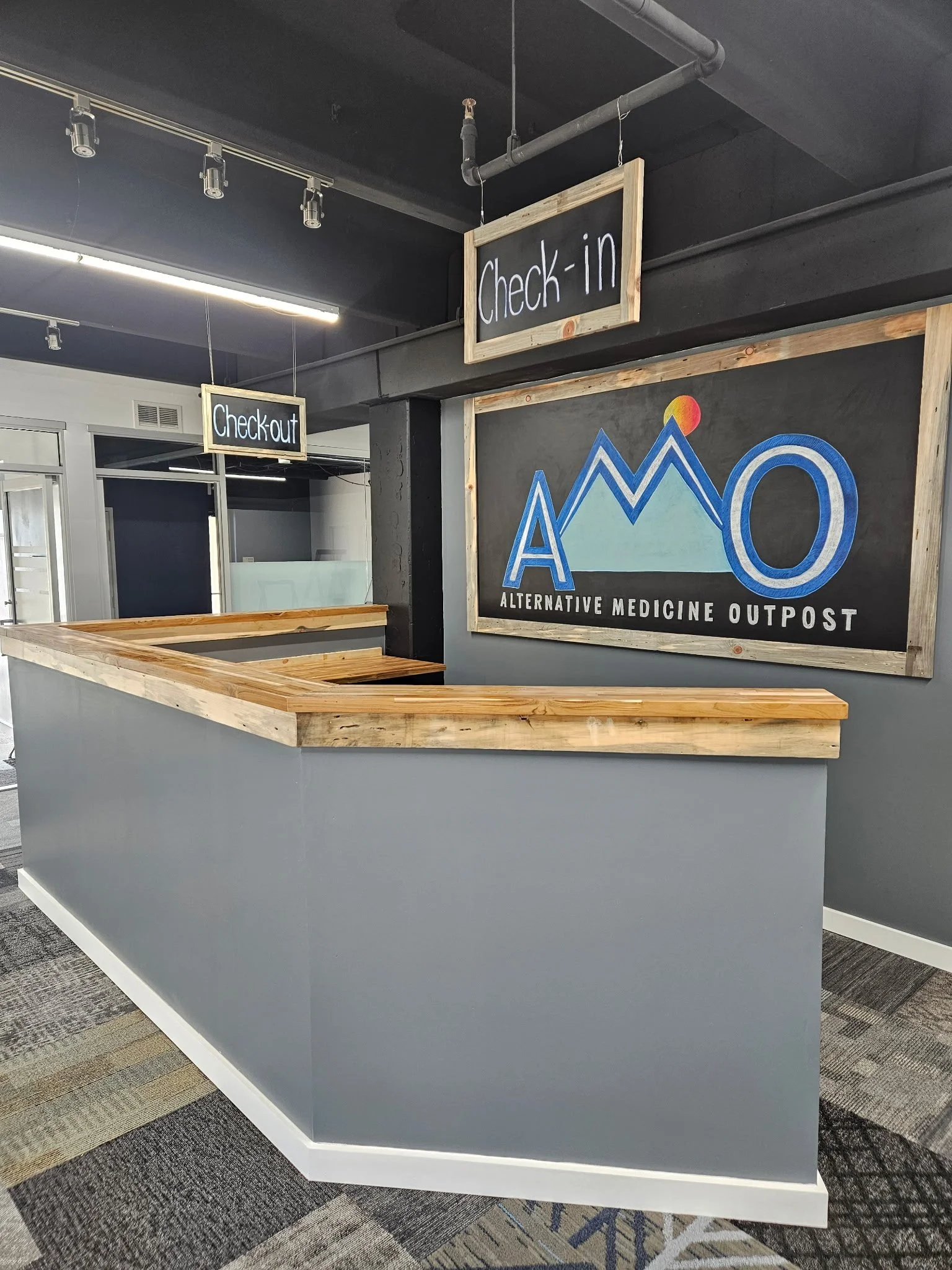 Reception desk at a modern medical outpost with signs for check-in and check-out, and a large logo for Alternative Medicine Outpost.