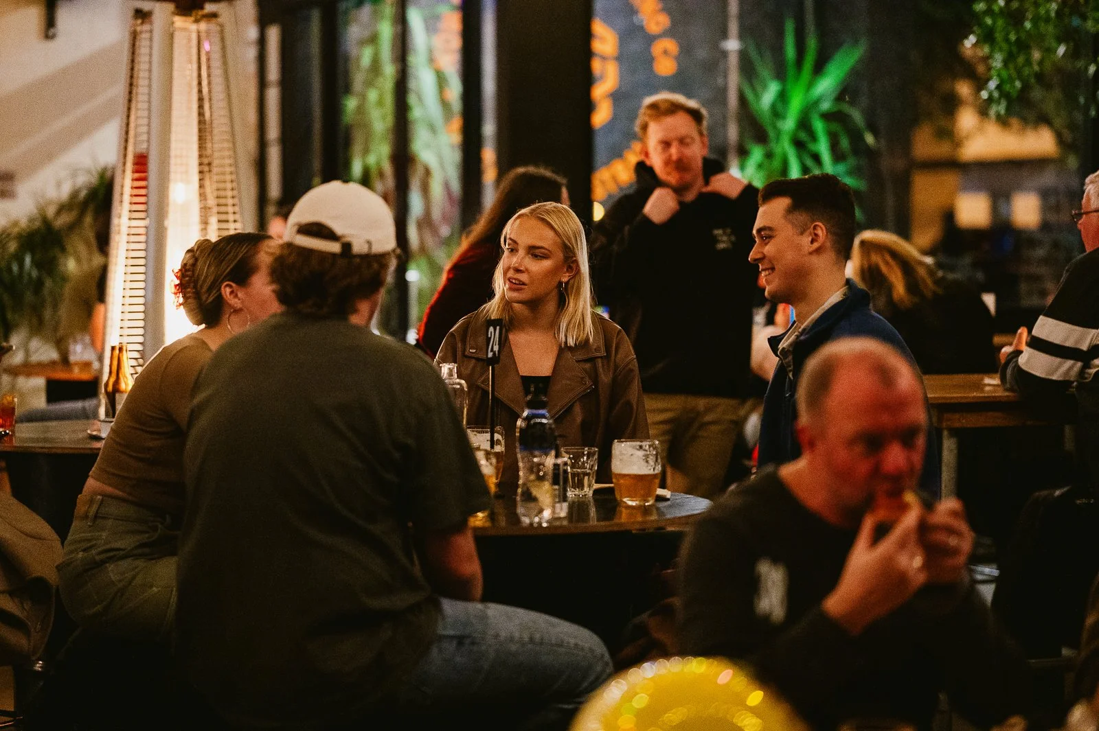 People gathered at a bar or pub, socializing and drinking beer, with neon and decorative lighting in the background.