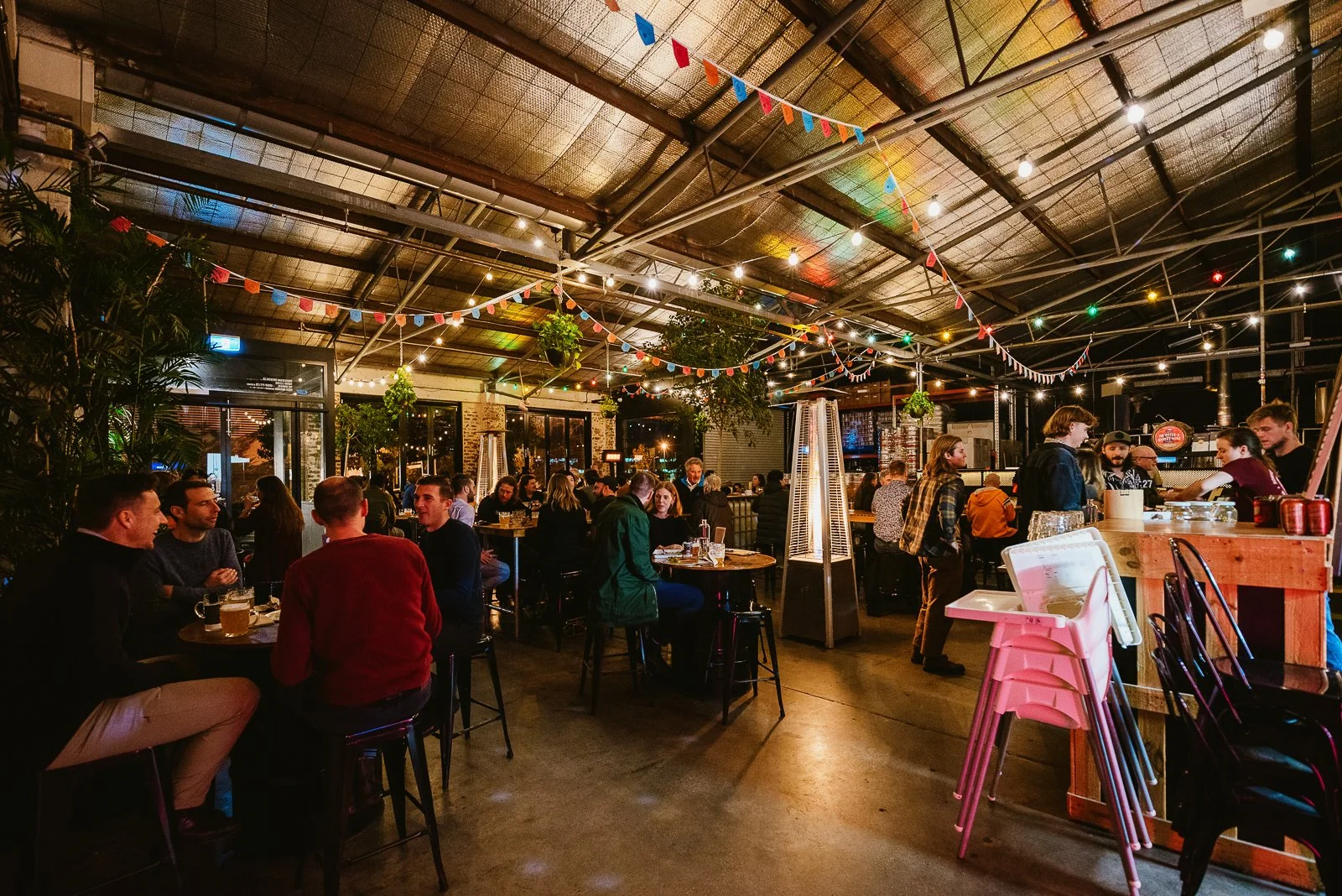 A lively indoor bar or restaurant with people socializing, colorful string lights and bunting decorations, heaters, and large windows at night.