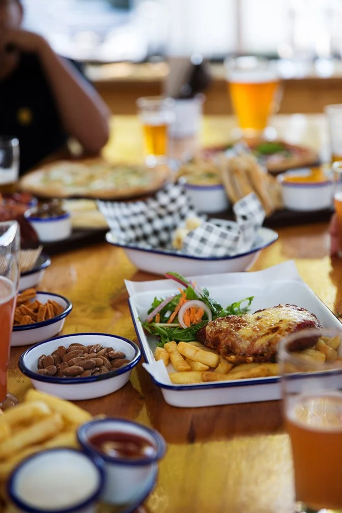 A table with various dishes including a plate of chicken with fries and salad, bowls of beans and sauces, and glasses of beer in a restaurant setting.
