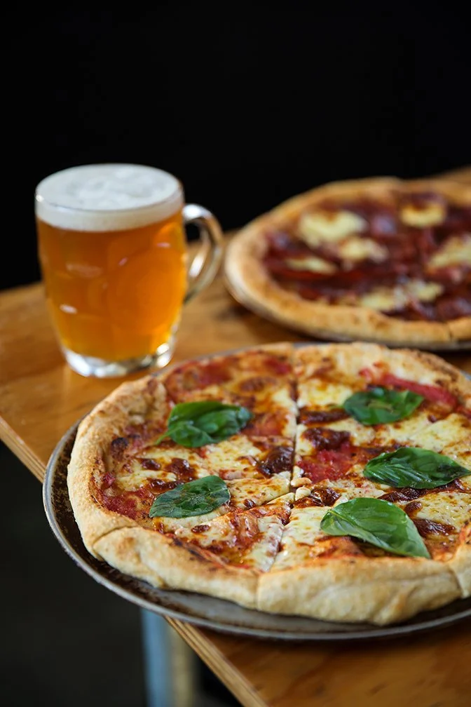 A pizza with cheese, tomato sauce, pepperoni, and basil leaves on a plate, another pepperoni pizza in the background, and a glass of beer on a wooden table.