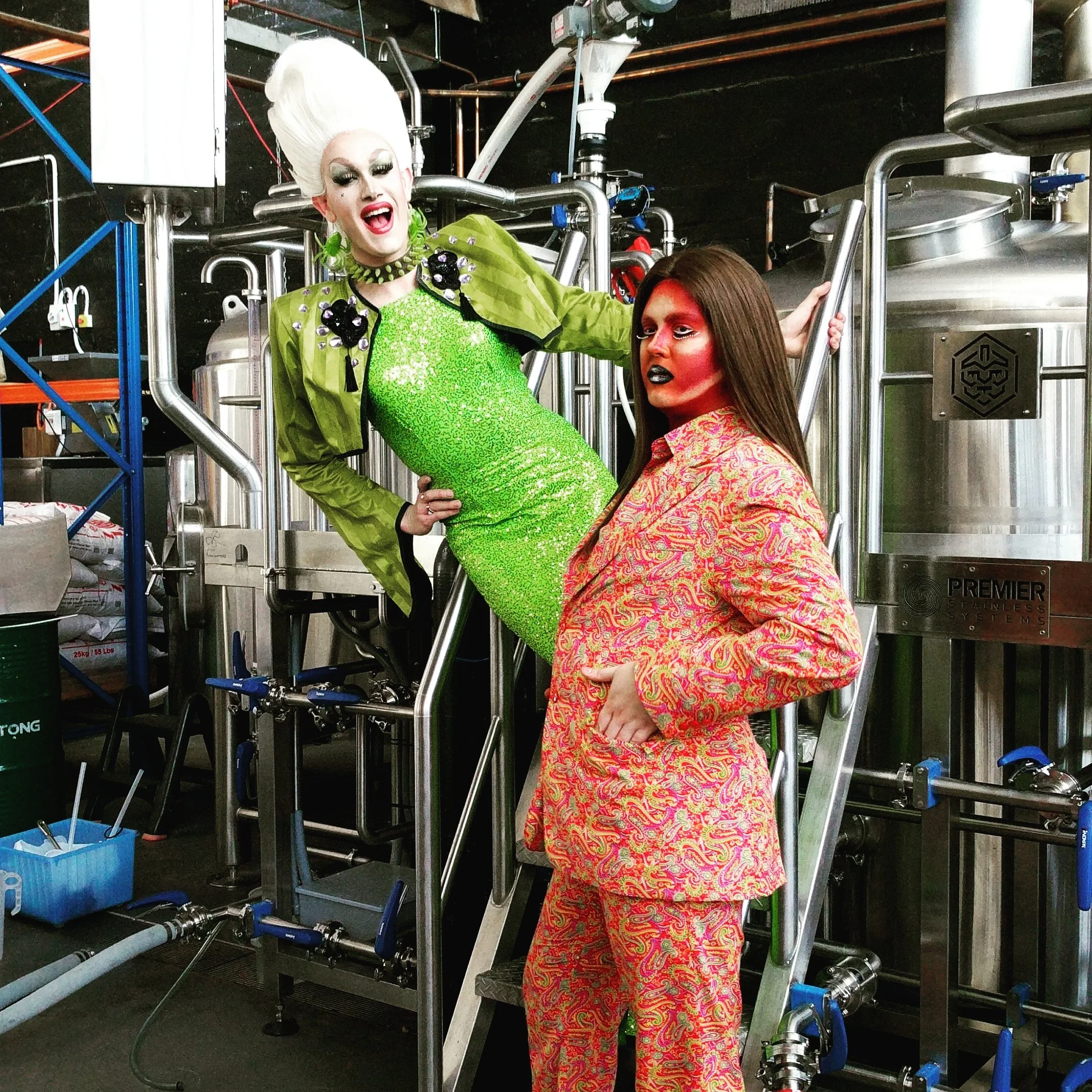 Two people dressed in bold, colorful outfits with dramatic makeup in an industrial setting with stainless steel equipment.