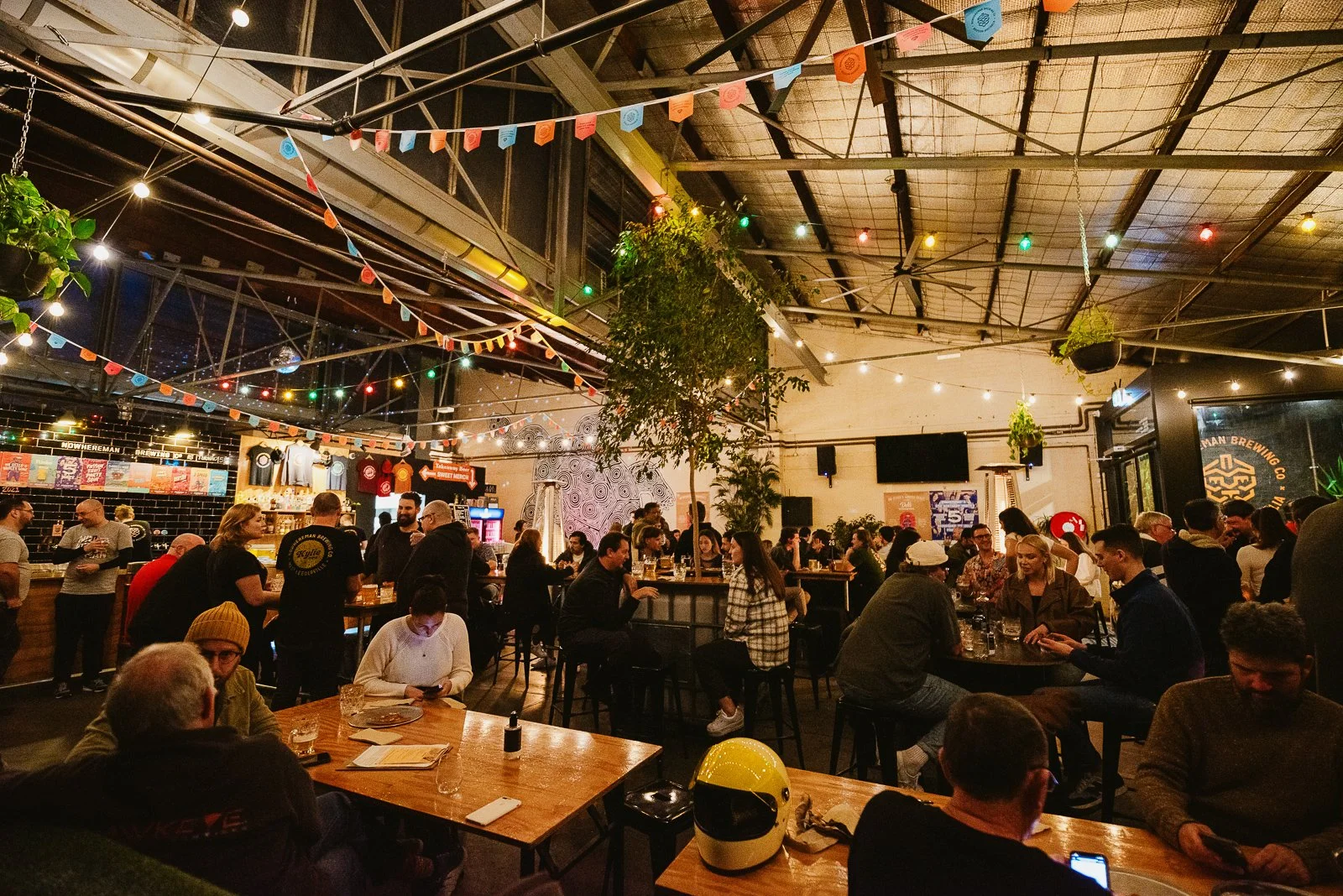 A bustling indoor bar and restaurant filled with people sitting at tables, socializing, and enjoying drinks. String lights and colorful pennant banners decorate the ceiling, with a large tree in the center of the space. The background features a bar 