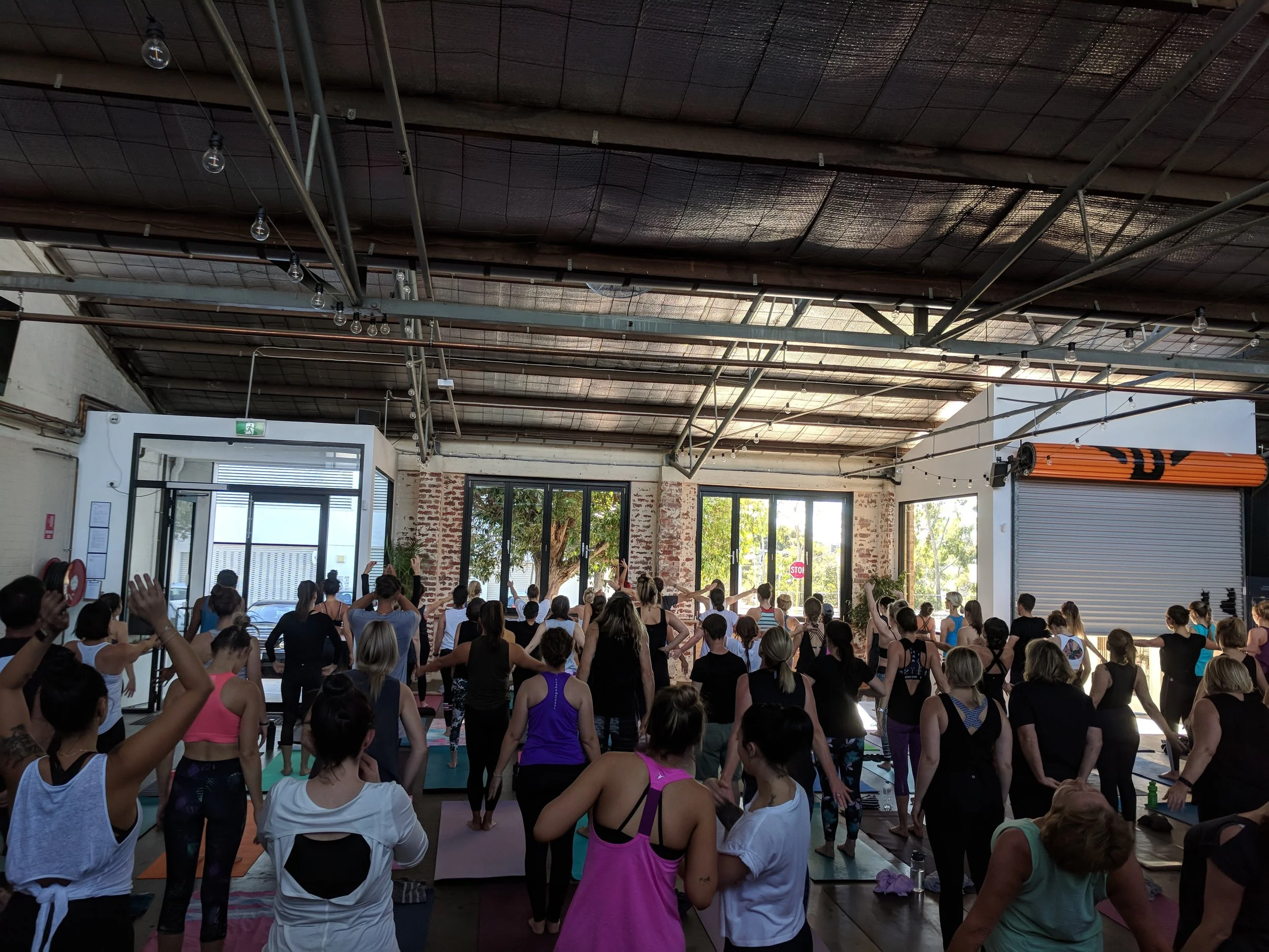 Group of people participating in a yoga class inside a spacious industrial-style studio with large windows and brick walls, some with hands raised.