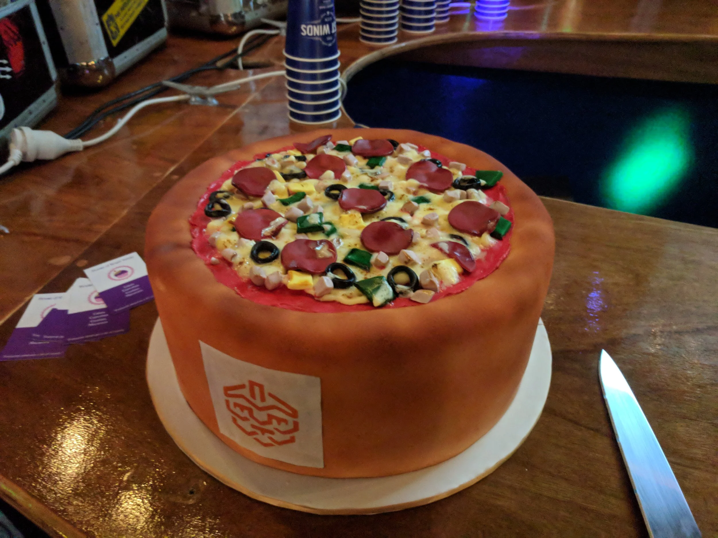 A cake designed to look like a pizza with toppings like pepperoni, black olives, green peppers, mushrooms, and cheese, placed on a wooden table.