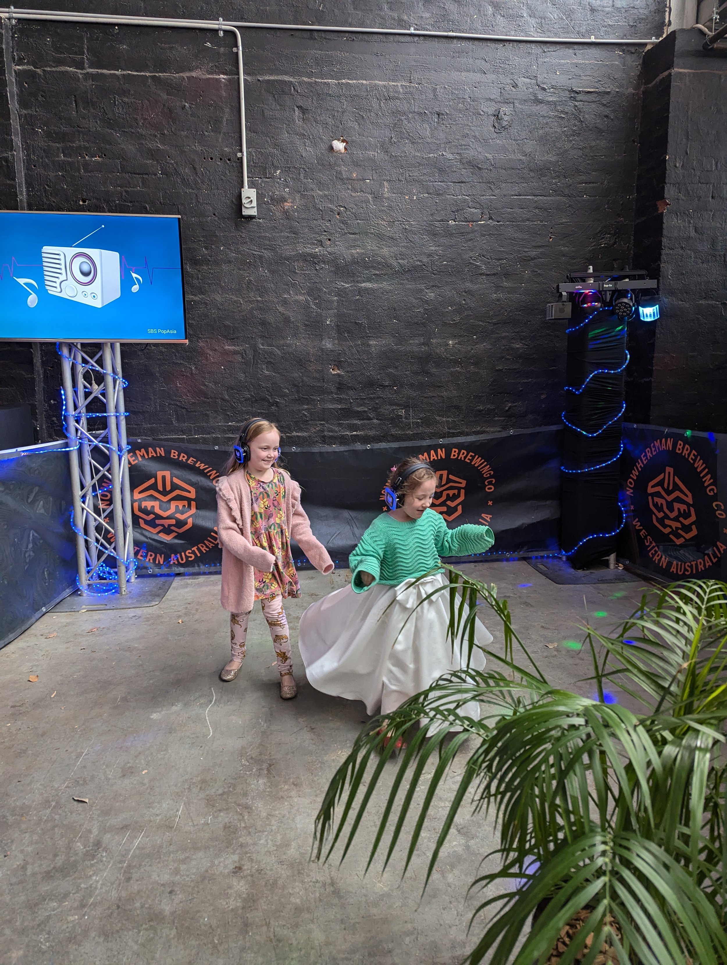 Two young girls wearing headphones are dancing and having fun at an indoor event at Nowerhemann Brewing in Western Australia. One girl is wearing a green sweater and a white skirt, while the other girl is wearing a pink cardigan and floral pants. A l
