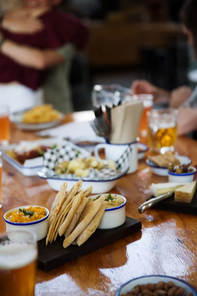 Close-up of a wooden table with various appetizers and drinks, including crackers with dips, small bowls of snacks, and glasses of beer in the background, with people standing around.