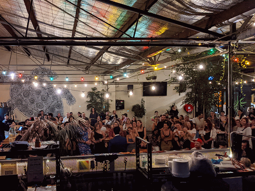 Crowd of people at a lively indoor event space with colorful string lights, a mural of a bear, and a corrugated metal ceiling.