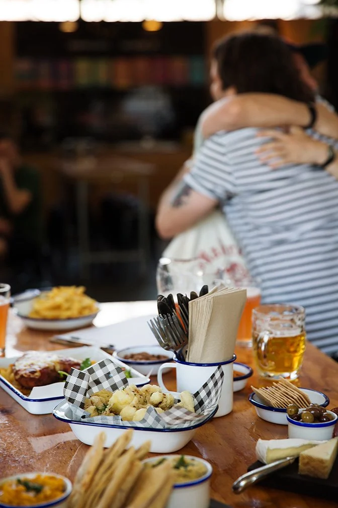 Table with various foods including fried snacks, cheese, olives, breadsticks, and drinks, with a person and a dog hugging in the background.