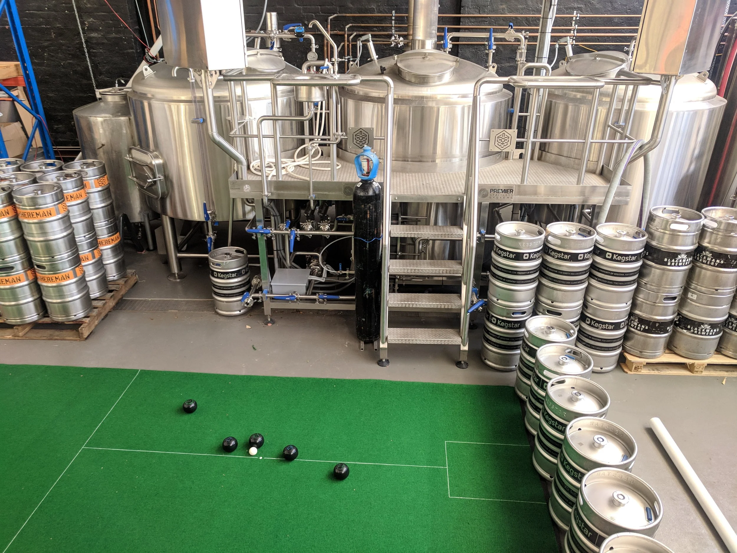 Beer brewing equipment with stainless steel fermentation tanks, beer kegs, and a small bocce ball game on a green mat in the foreground.