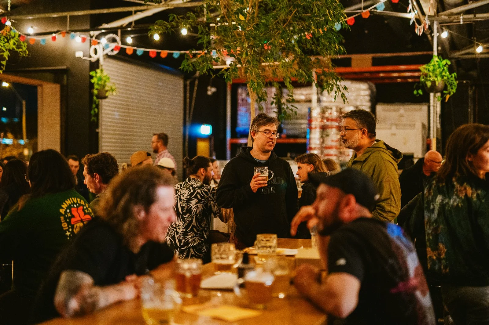 People socializing indoors at a bar or brewery, with a long wooden table and drinks in front of them. String lights and hanging plants decorate the space, with a lively atmosphere.