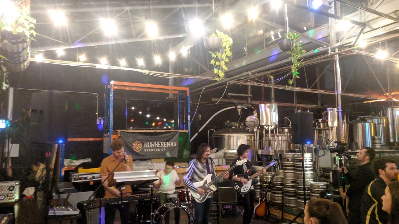 A band performing live in a brewery with brewing equipment and shelves of beer kegs in the background, under bright string lights.