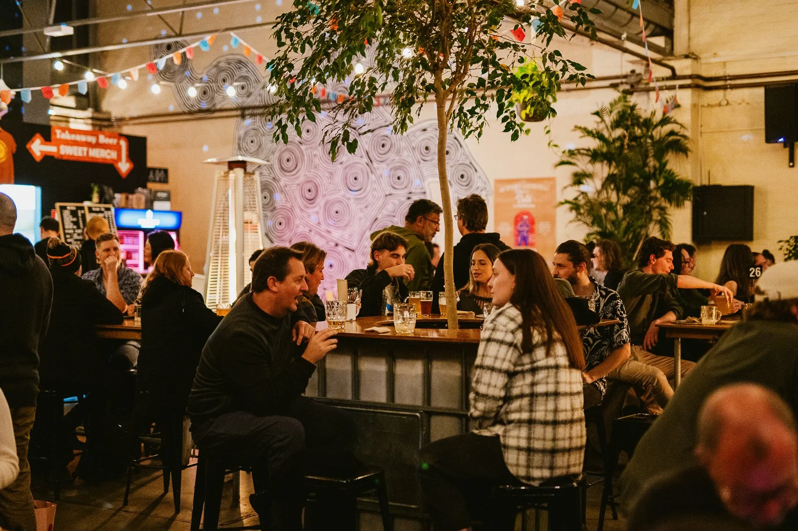 People sitting at long communal tables in a lively bar or restaurant with a tree in the center, colorful decorations, and dim ambient lighting.