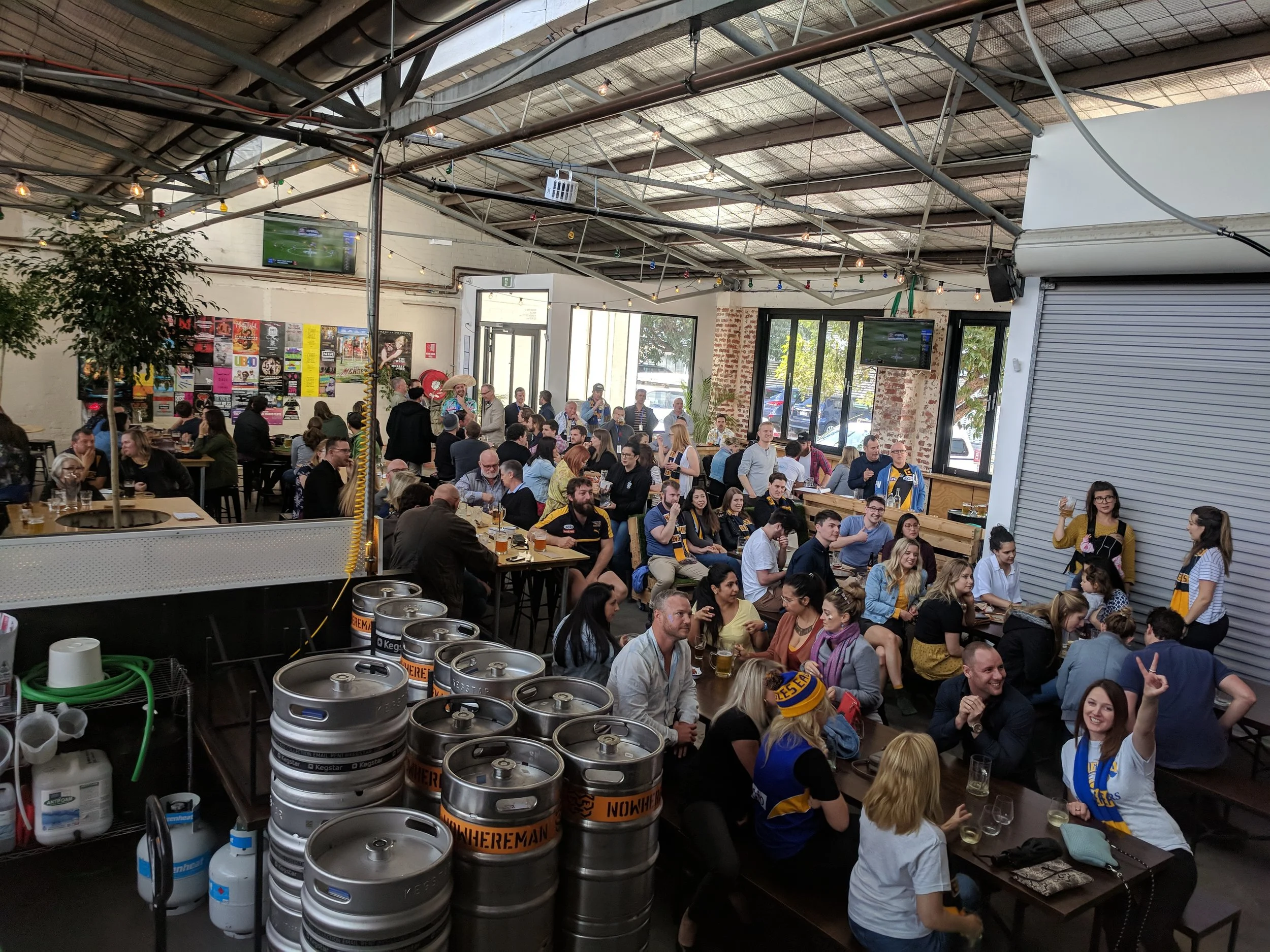 Crowd of people gathered in a brewery or pub with beer kegs in the foreground and multiple TVs on the walls watching a sports game.