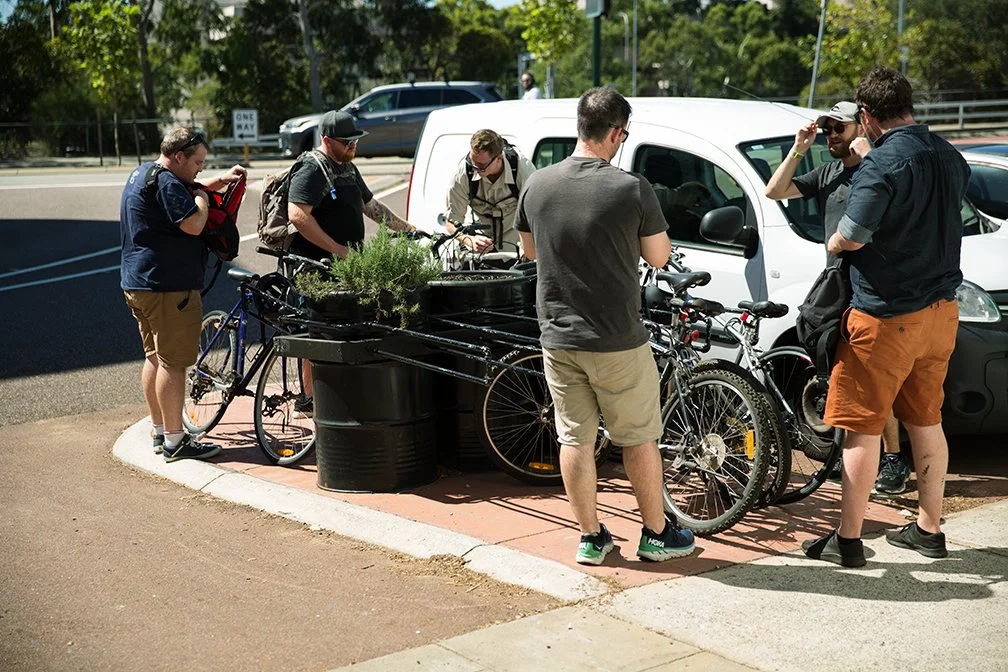 Six people gathered around parked bicycles near a white van in an outdoor area, with some holding or adjusting the bikes and others talking. Trees and vehicles are in the background.