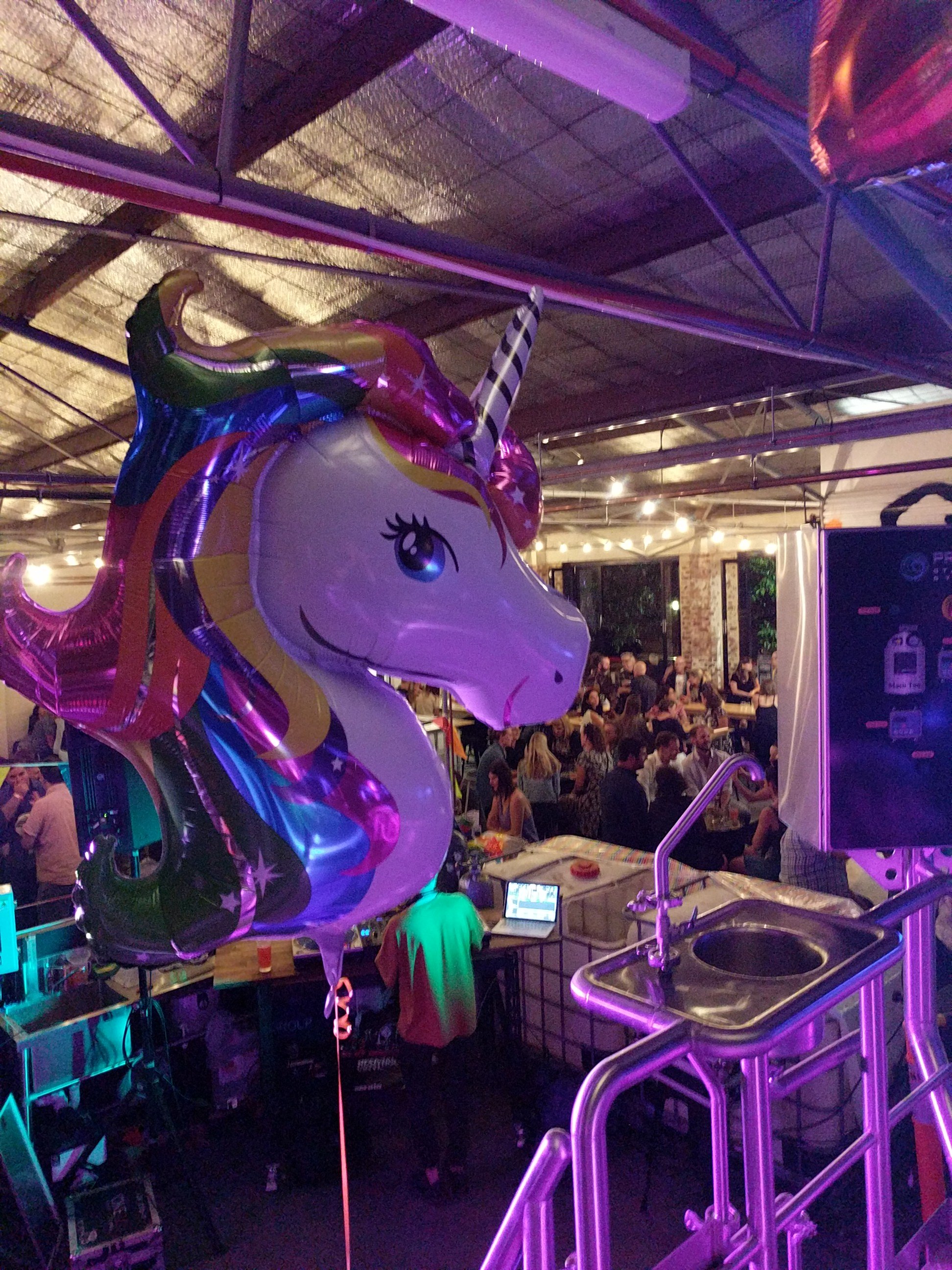 Unicorn-shaped balloon with rainbow colors in a dimly lit indoor setting, possibly a party or celebration, with a crowd of people in the background.